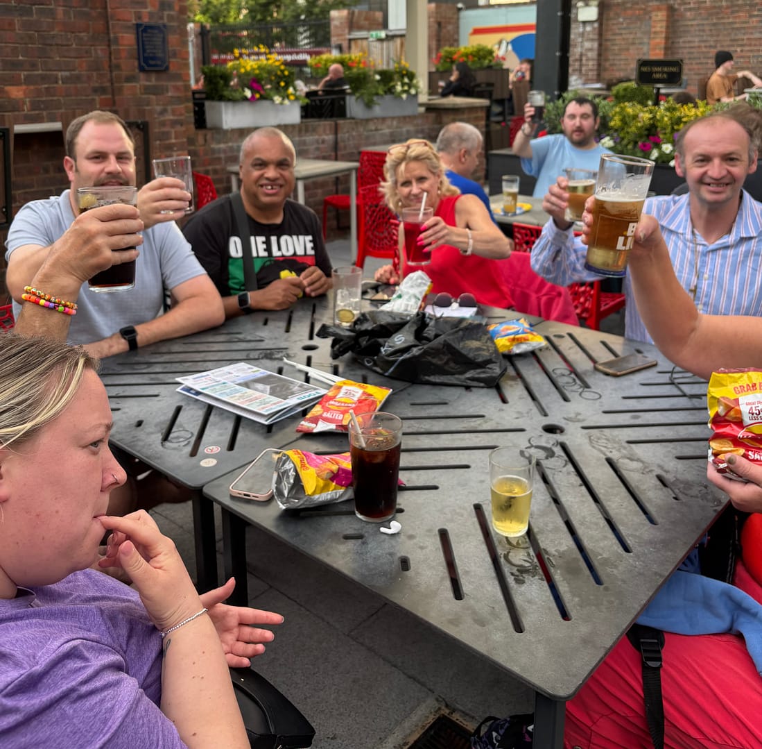 A group of people are sitting around a table in a pub garden raising drinks.