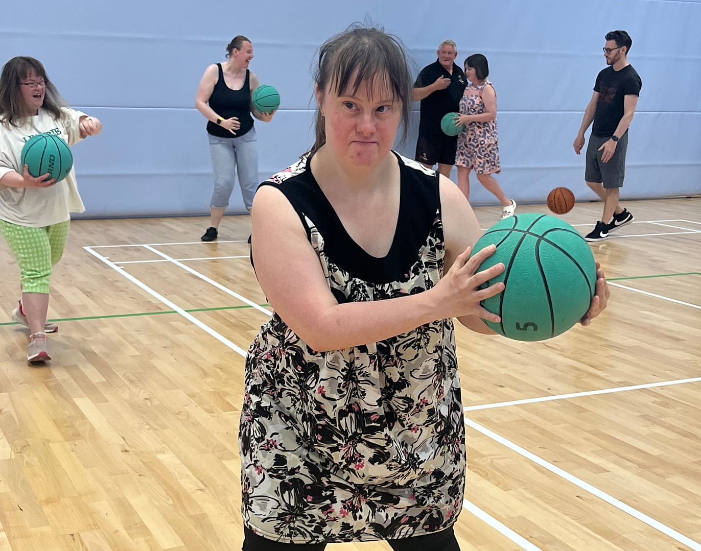 A young woman is in a gym holding a green basketball in her hands. Other people in the background are also holding balls.