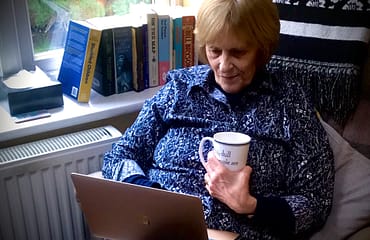 A woman sits in an armchair with a cup of tea while browsing on a laptop. A selection of books sit on a windowsill beside her.