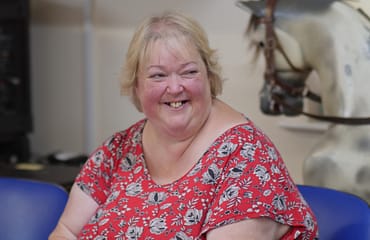 A smiling blonde woman is wearing a red top with black and white flowers. She is sitting on a blue chair. The head of a rocking horse can be seen in the background.