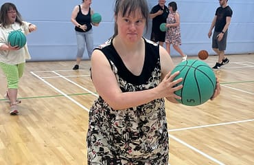 A young woman is in a gym holding a green basketball in her hands. Other people in the background are also holding balls.