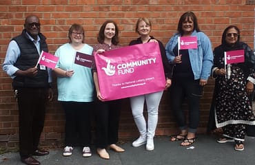Five women and one man are standing holding a pink National Lottery Banner and waving National Lottery flags