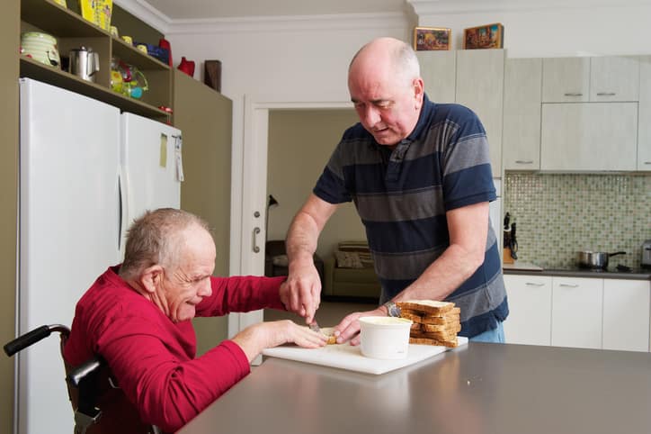 An older man is helping a disabled man in a wheelchair eat a meal.