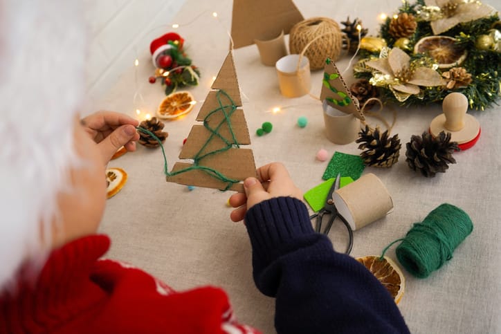 Cardboard cut in the shape of a Christmas tree. Green wool is being threaded around it. Other craft materials are on the table.