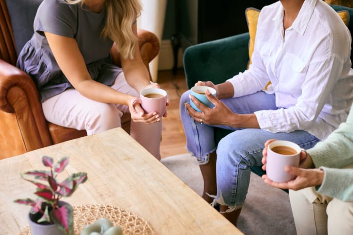 Three women are sitting around a table holding mugs of tea.